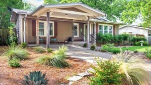A single-story house with a covered porch, a front yard with various plants, and a clear sky.