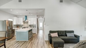 A spacious living area with a dark grey sofa, a wooden floor, and a kitchen with white cabinets and a blue island.