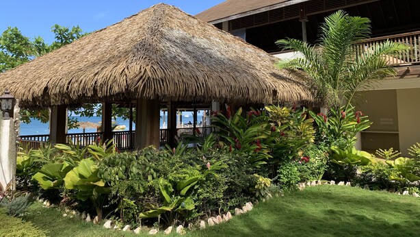 A thatched-roof structure with a view of the sea, surrounded by lush tropical plants and a well-maintained lawn. - Family Comfort In Jamaica - Enjoy 7 Miles Of White Sand Beach! Villa by RedAwning (Negril)