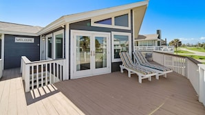 Uma casa moderna com um letreiro 'WELCOME', um deck com duas poltronas listradas e uma porta de vidro que dá para um alpendre coberto.