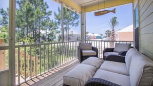 A covered outdoor area with a wicker sofa and chairs, a wooden deck, and a view of trees and buildings. - The Rookery Iii 7011 3 Bedroom Cottage by RedAwning (Gulf Shores)