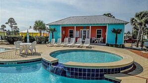 A pool area with a hot tub, lounge chairs, and a small building with a red and blue facade. - The Rookery Iii 7011 3 Bedroom Cottage by RedAwning (Gulf Shores)