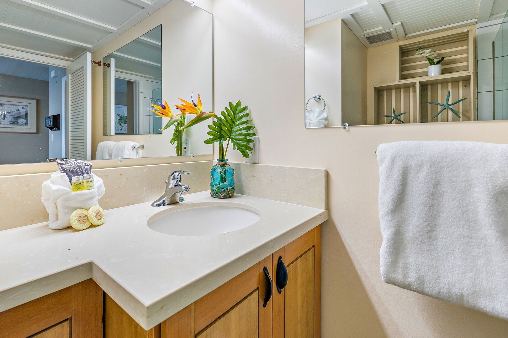 Un bagno con un lavandino bianco, mobili impiallacciati in legno, uno specchio grande e oggetti decorativi tra cui stelle marine e una pianta.