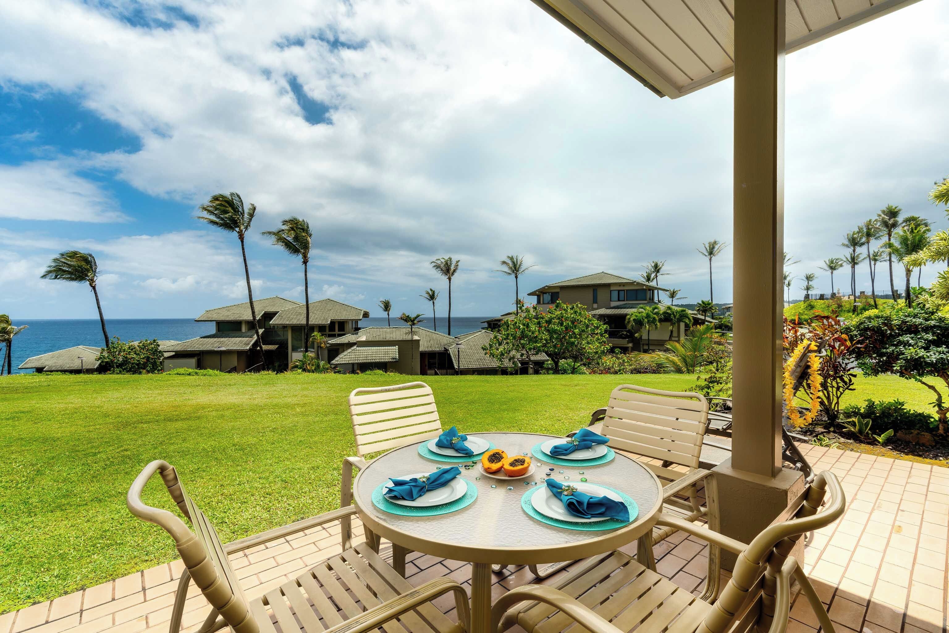 Une table ronde entourée de quatre chaises, un jardin tropical et une vue sur l’océan et les maisons qui se dessinent à l’arrière-plan.
