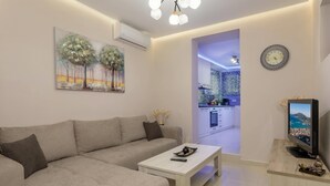 A modern living room with a sofa, coffee table, and a TV. There is a painting on the wall and a kitchen visible through an open doorway. - Evia's Apartment   New Apartment In Town! (Zakynthos)