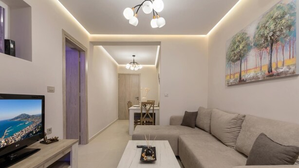 A modern living room with a flat-screen TV, a white coffee table, a beige sofa, and a dining area with a table and chairs. - Evia's Apartment   New Apartment In Town! (Zakynthos)