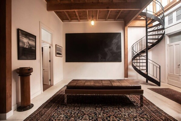 A spacious room with a large black artwork on the wall, a wooden bench, a patterned rug, and a spiral staircase. - w* | Villa with Rooftop Terrace in Barranco (Barranco)