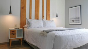 Un lit bien fait, avec du linge de lit blanc et un couvre-lit à motifs, une petite table de chevet en bois avec un tiroir, et un panneau en bois fixé au mur.