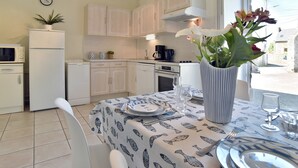 A kitchen with white cabinets, a microwave, a refrigerator, and a dining table set with plates, glasses, and a vase of flowers. - L'Armoricaine (Cancale)