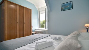 A bedroom with a bed, a wooden wardrobe, a window with a view, and a painting on the wall. - L'Armoricaine (Cancale)
