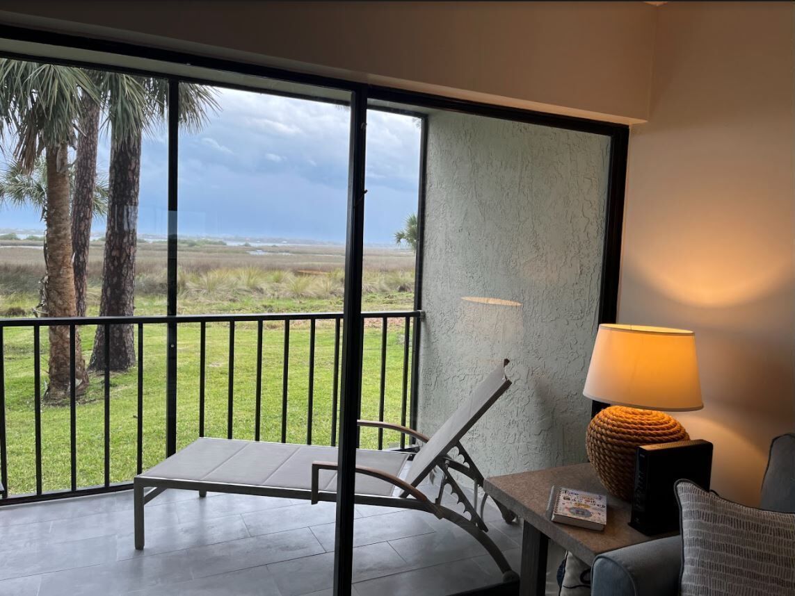 A balcony with a view of the beach, a chair, a table, and a lamp.