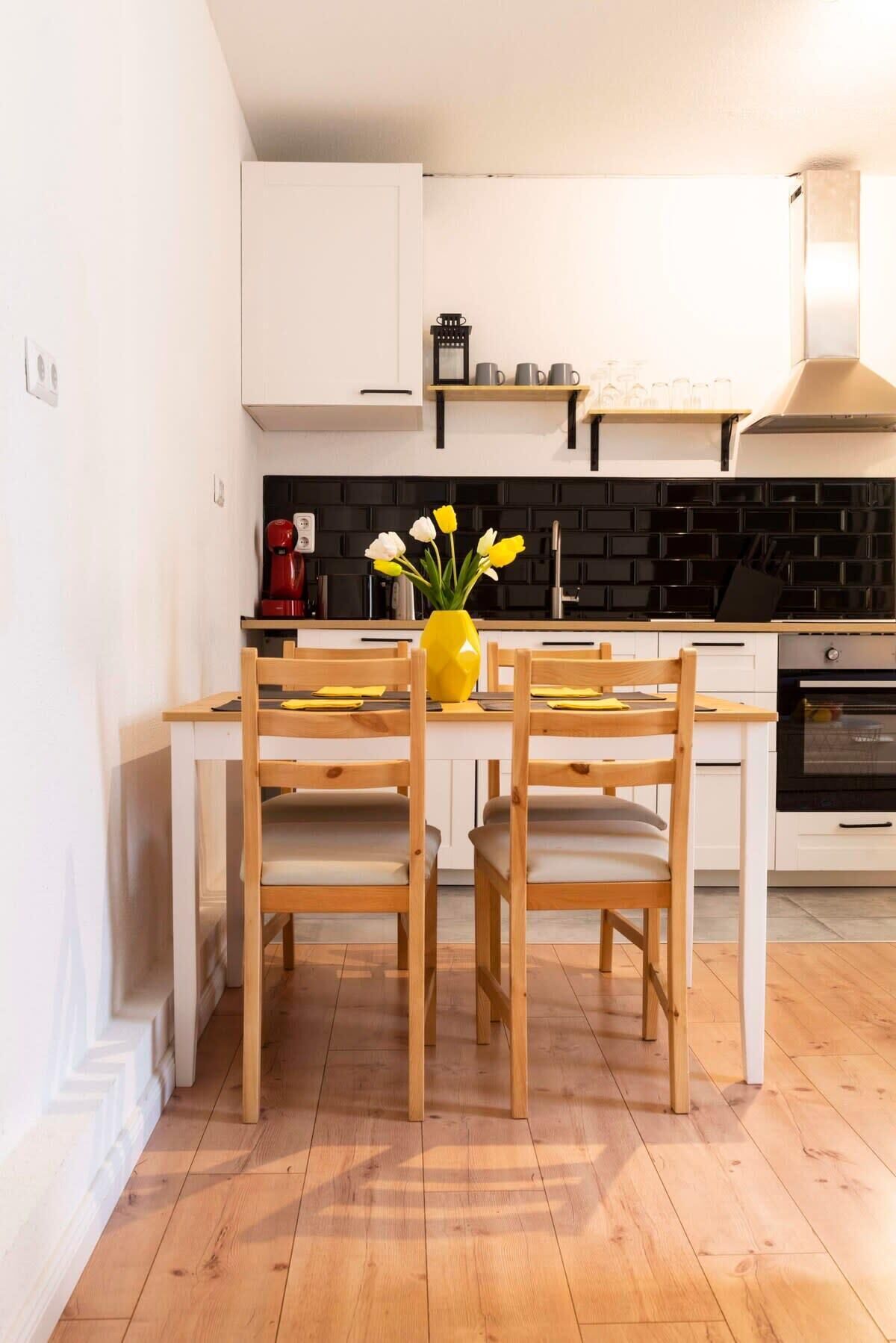 Une cuisine moderne avec des armoires blanches, un plan de travail noir et un revêtement de sol en bois. On y trouve une table à manger avec deux chaises et un vase rempli de fleurs.