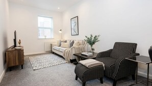 Una sala de estar con sofá, sillón, otomanas, una mesita auxiliar y una pequeña mesa con lámpara.