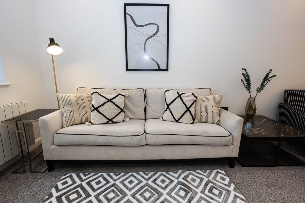 A living room with a sofa, patterned cushions, a framed abstract artwork, a floor lamp, a small table, and a vase with green foliage.
