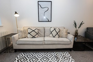A living room with a sofa, patterned cushions, a framed abstract artwork, a floor lamp, a small table, and a vase with green foliage.