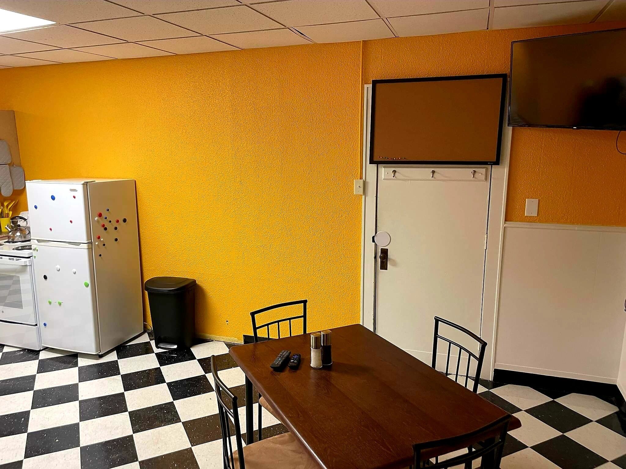 A room with a checkered floor, a wooden table with chairs, a refrigerator with magnets, a trash bin, and a door with a bulletin board.