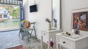 A modern room with a large window overlooking a body of water, a chair with a hat, a desk with a mirror, and a painting on the wall. - ECO SIRMA FLARIS (Milos)
