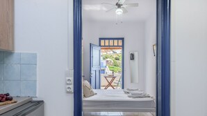 A narrow hallway leading to a bedroom with a bed, a small table, and a chair. There is a ceiling fan above the bed and a view of a balcony with a table and chairs outside. - ECO SIRMA FLARIS (Milos)