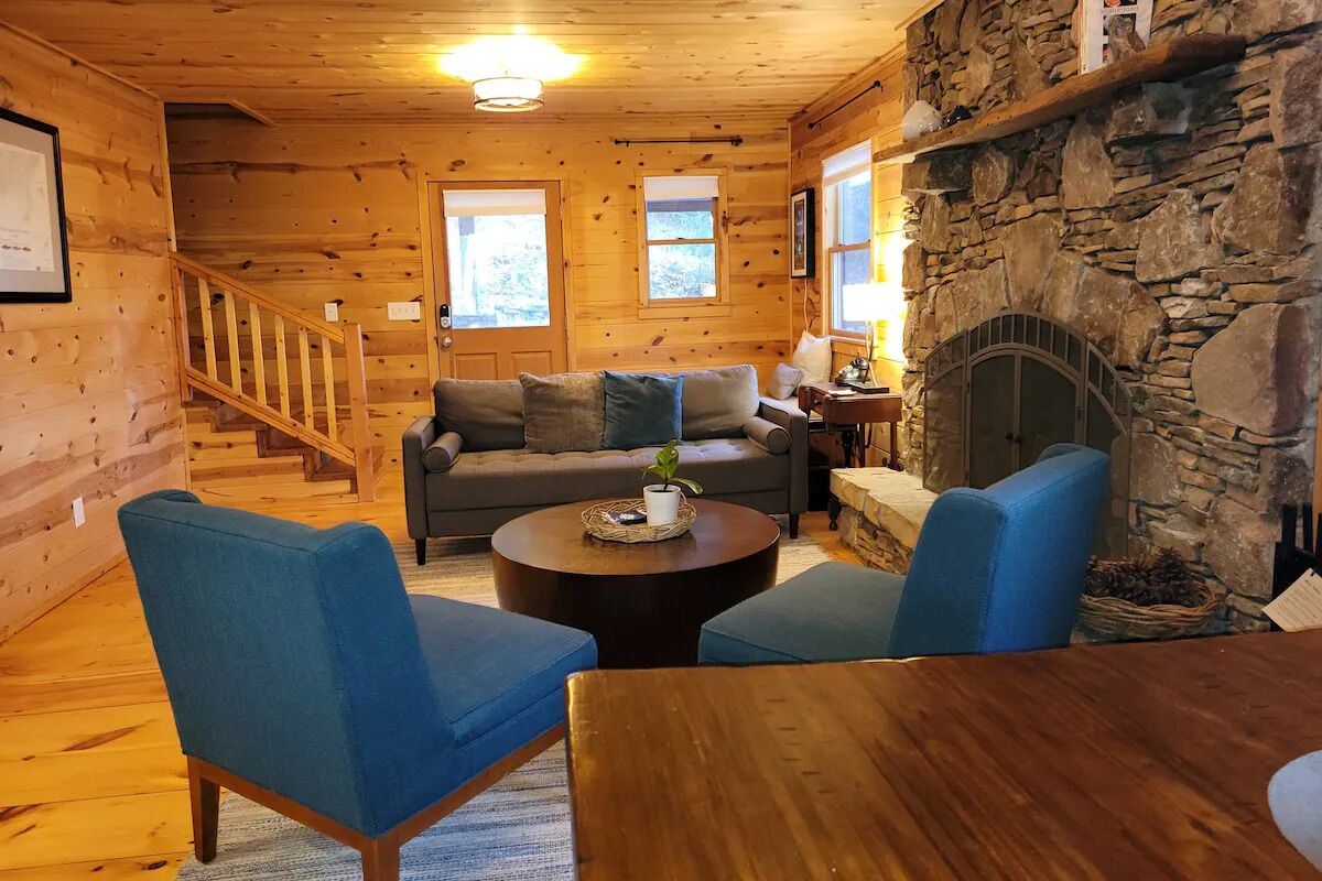 Interior acogedor de una cabaña con chimenea de piedra, paredes de madera, un sofá y sillones azules.