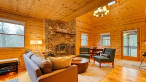 Interior acogedor de una cabaña de troncos con chimenea de piedra, muebles de madera y ventanas amplias.