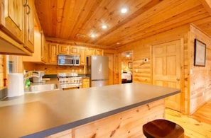 Interior de uma cabana de madeira com cozinha, armários de madeira, bancada de aço inoxidável e geladeira.