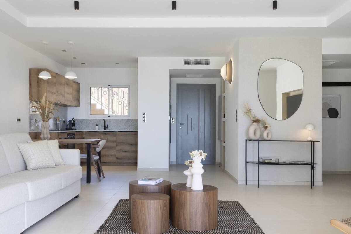 A modern living room with a sofa, coffee table, and a dining area with chairs. There is a kitchen in the background with wooden cabinets and a window. A mirror is on the wall, and there is a console table with a lamp.