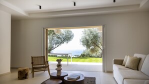 Una sala moderna con un sofá, una silla de madera, una mesita con un florero y vista a un paisaje verde a través de una puerta de vidrio.