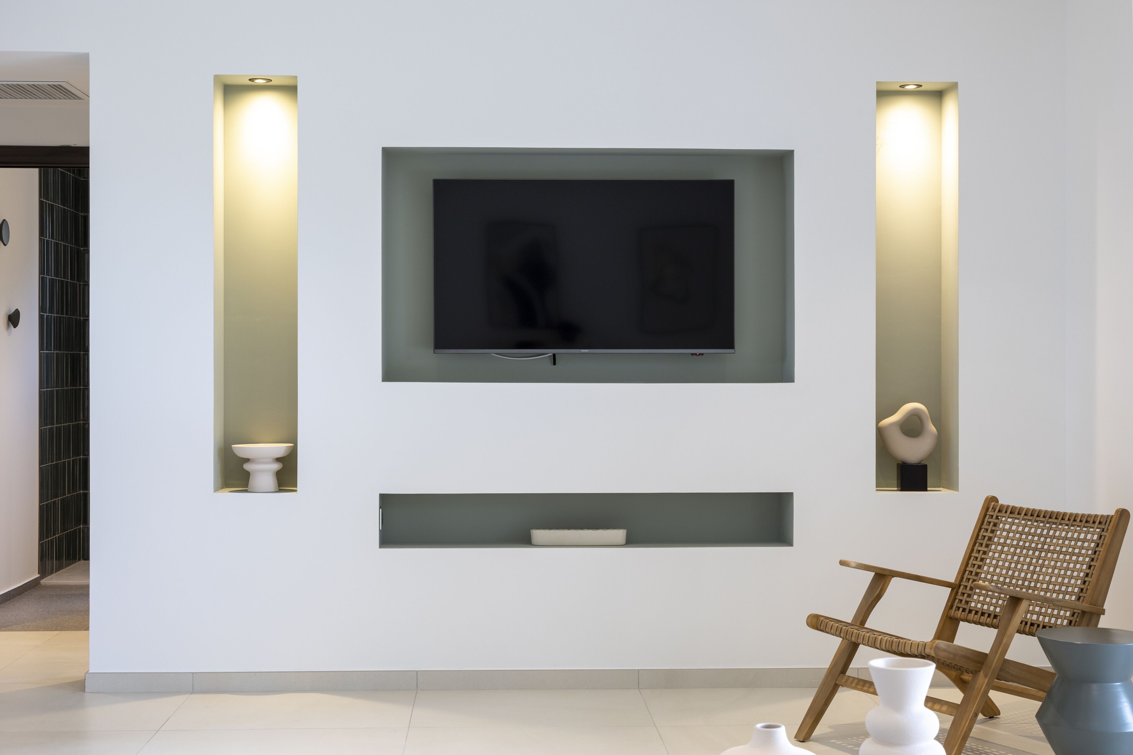A modern living room with a flat-screen TV mounted on the wall, flanked by two wall-mounted sconces. There is a wooden chair and a small white table in the room.