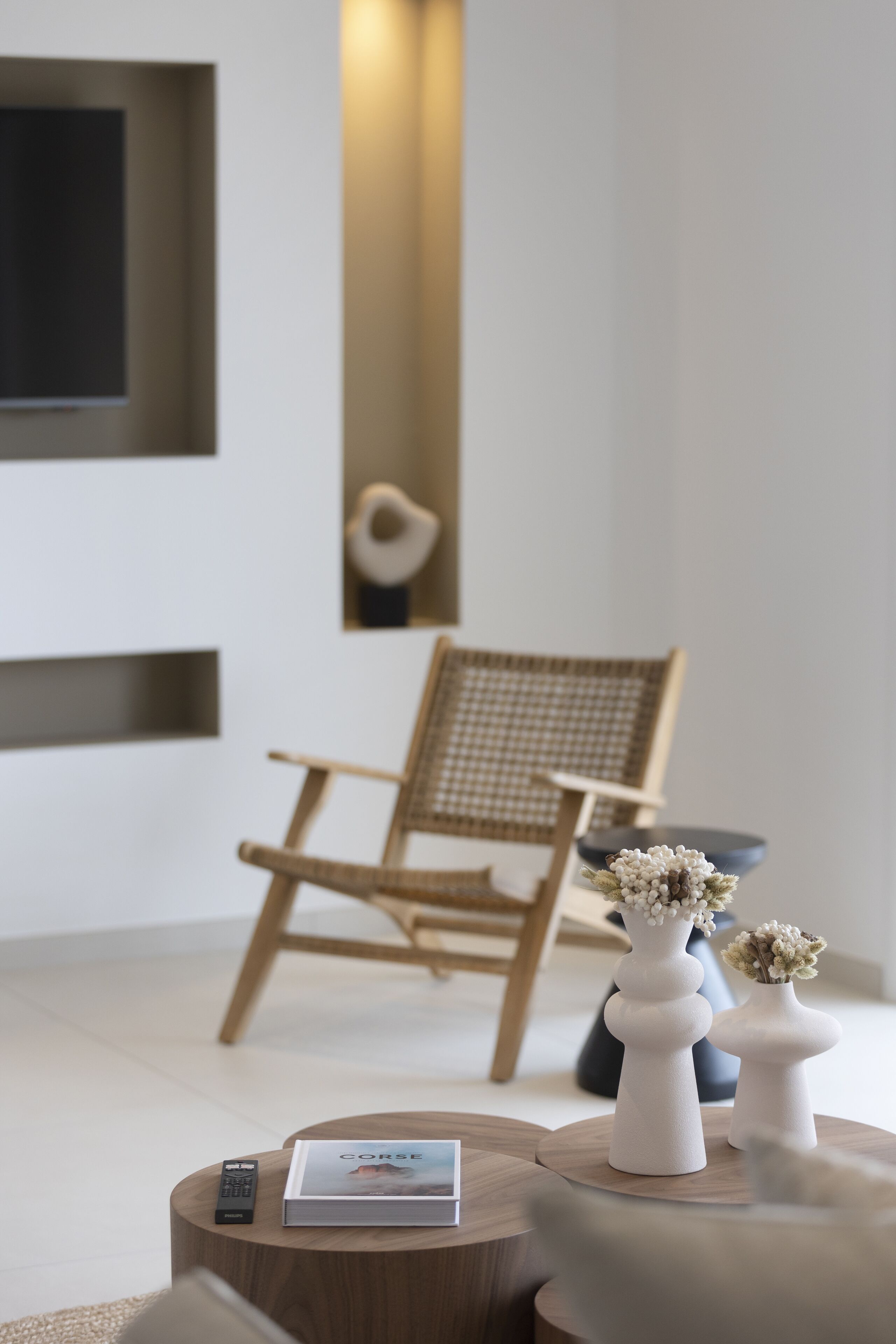 A modern living room with a wooden chair, two white vases with flowers, a remote control, and a book titled 'CORSE'.