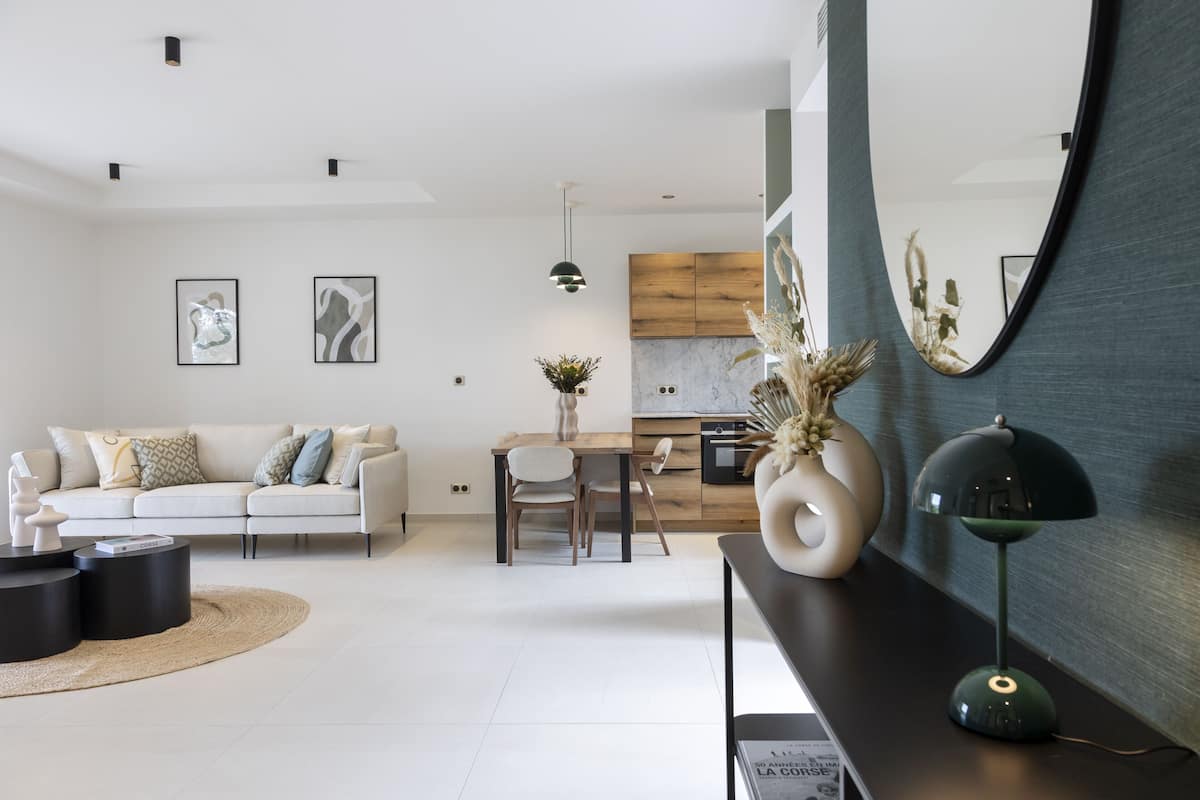 A modern living room with a sofa, coffee table, and a dining area with chairs. There is a kitchen in the background with wooden cabinets and a marble countertop.