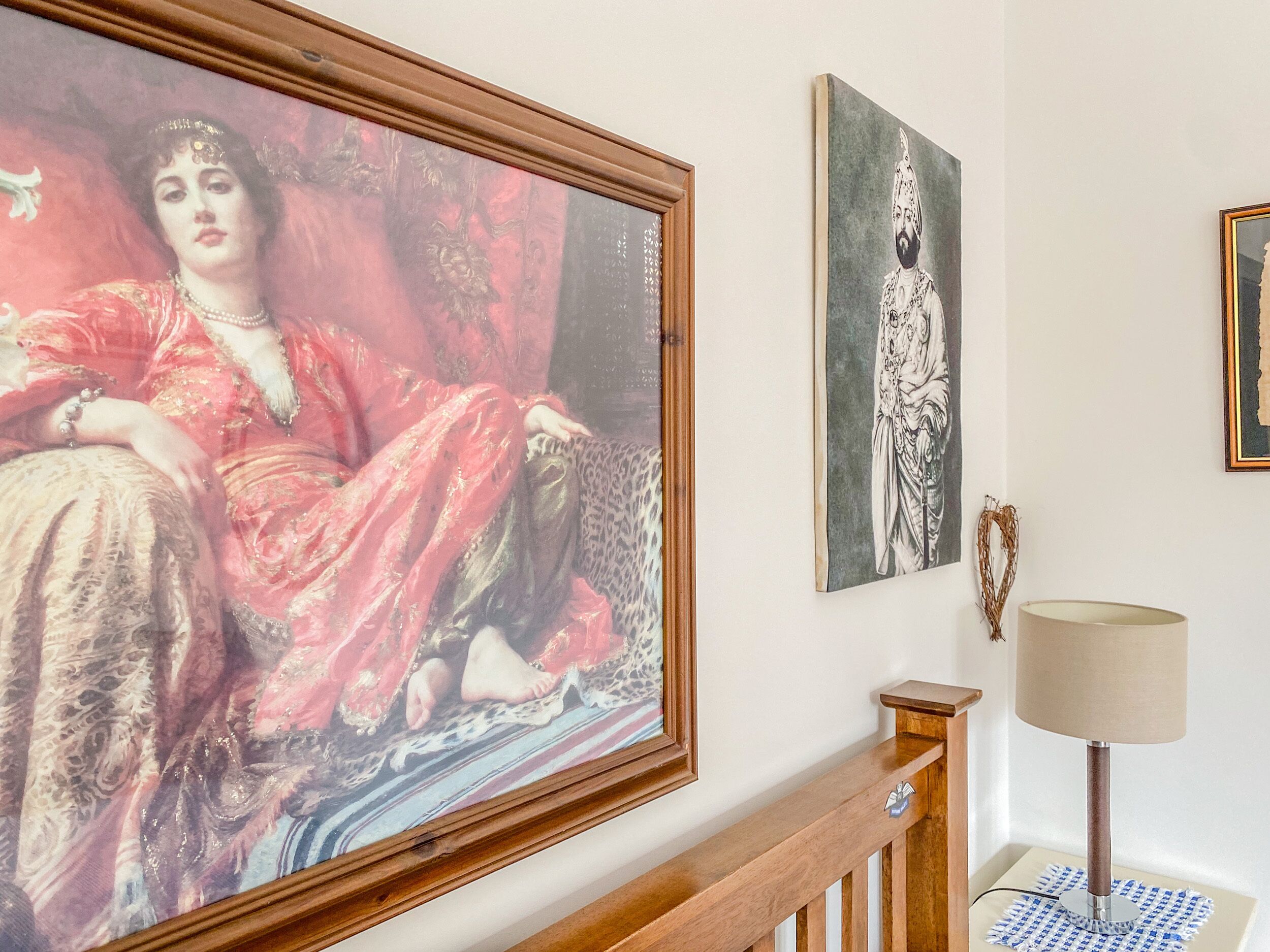 A hallway with a large framed painting of a woman in a red dress, a smaller framed portrait, and a lamp on a side table.