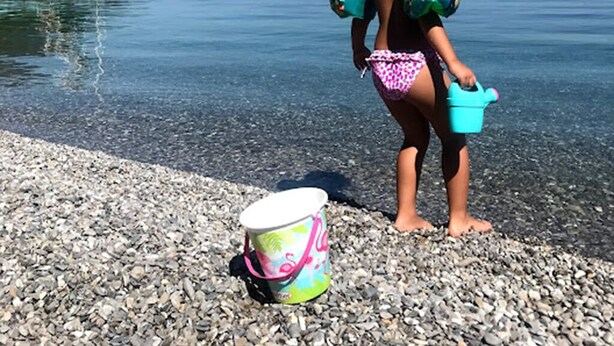 Una persona de pie en una playa pedregosa con vista despejada a una masa de agua tranquila y una isla distante.
