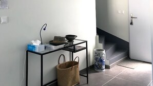 Un pasillo con una puerta blanca, una escalera, un cuadro enmarco, una mesita con una cesta y una lámpara de pie.