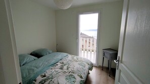 A bedroom with a bed, a small desk, and a door leading to a balcony with a view of buildings and a body of water.