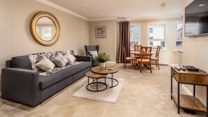 A living room with a grey sofa, a round mirror, a coffee table, and a dining area with wooden chairs and a table. - London Gate No1 - Donnini Apartments (Kilmarnock)