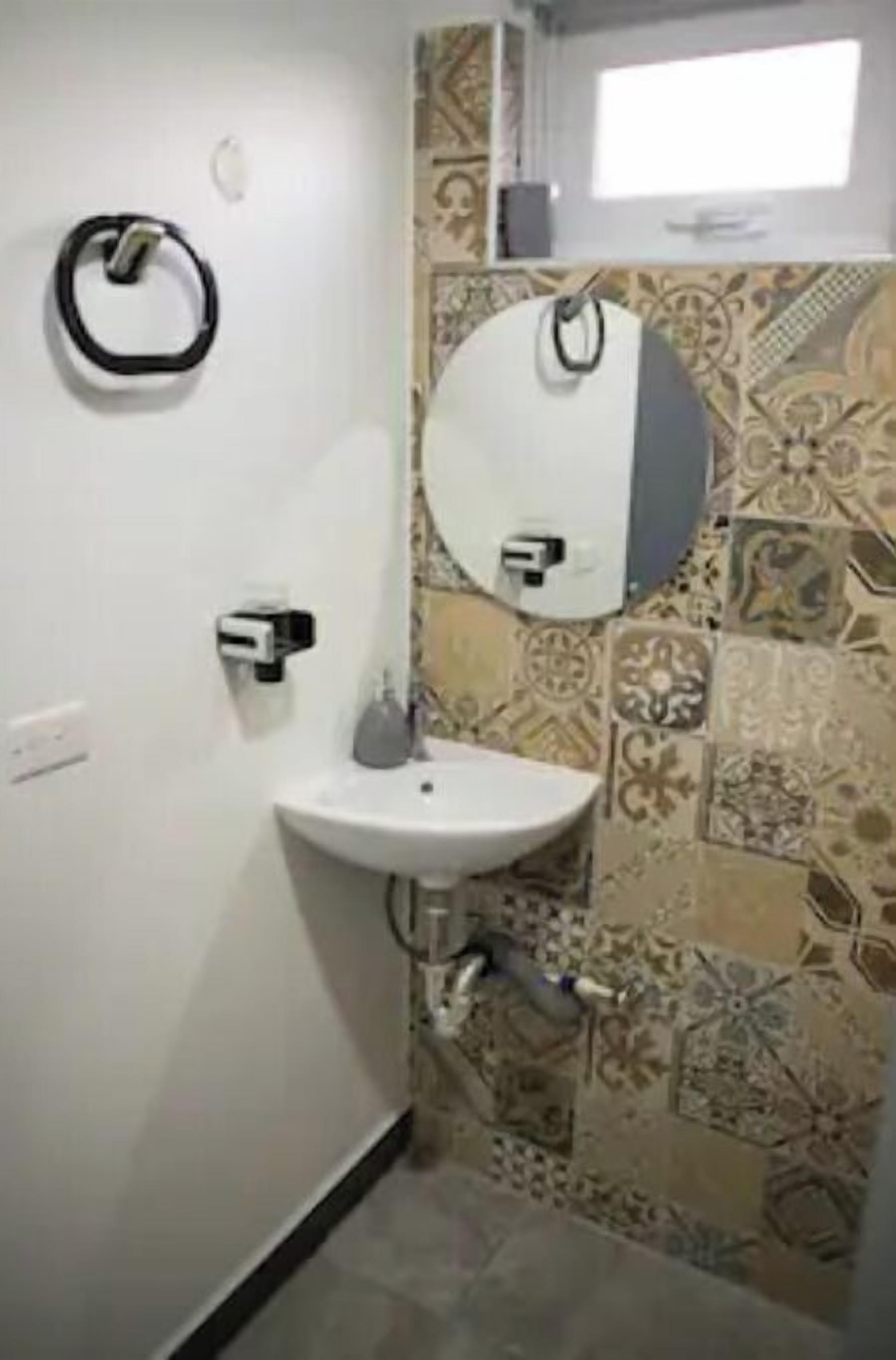 A bathroom with a white sink, a towel dispenser, and a mirror. The walls have patterned tiles.