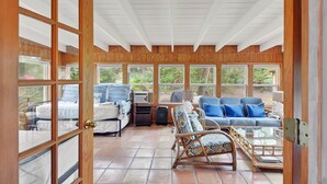 A sunroom with a wooden ceiling, large windows, a sofa, chairs, and a bed.