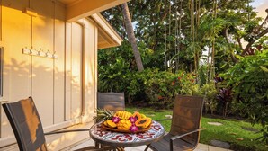 Un patio con dos sillas y una mesa, un jardín tropical al fondo y un edificio de fachada blanca.