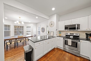 Una cocina moderna con armarios blancos, una isla central, electrodomésticos de acero inoxidable y una zona de comedor con mesa y sillas de madera.