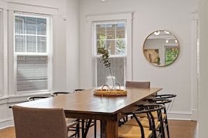 A dining area with a wooden table, chairs, a vase with greenery, and a round mirror on the wall.