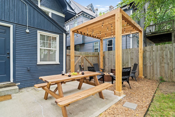 A wooden pergola with string lights, a picnic table with benches, and a fire pit in a backyard.