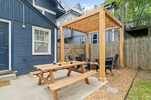 Una pérgola de madera con luces colgantes, una mesa de picnic con bancos y un brasero en un patio trasero.
