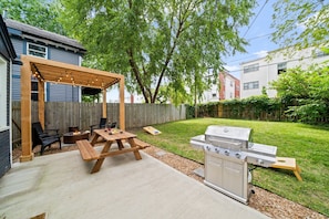 A backyard with a wooden pergola, picnic table, barbecue grill, and a fenced area.