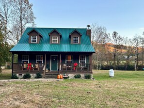 A two-story house with a green metal roof, a front porch with a railing, and a small front yard with pumpkins and a Christmas wreath.