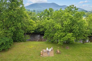 A grassy outdoor area with a fire pit and seating, surrounded by dense trees and a mountain in the background. - Riverside Cabin w/Fishing, Hot Tub & Firepit (COSBY)