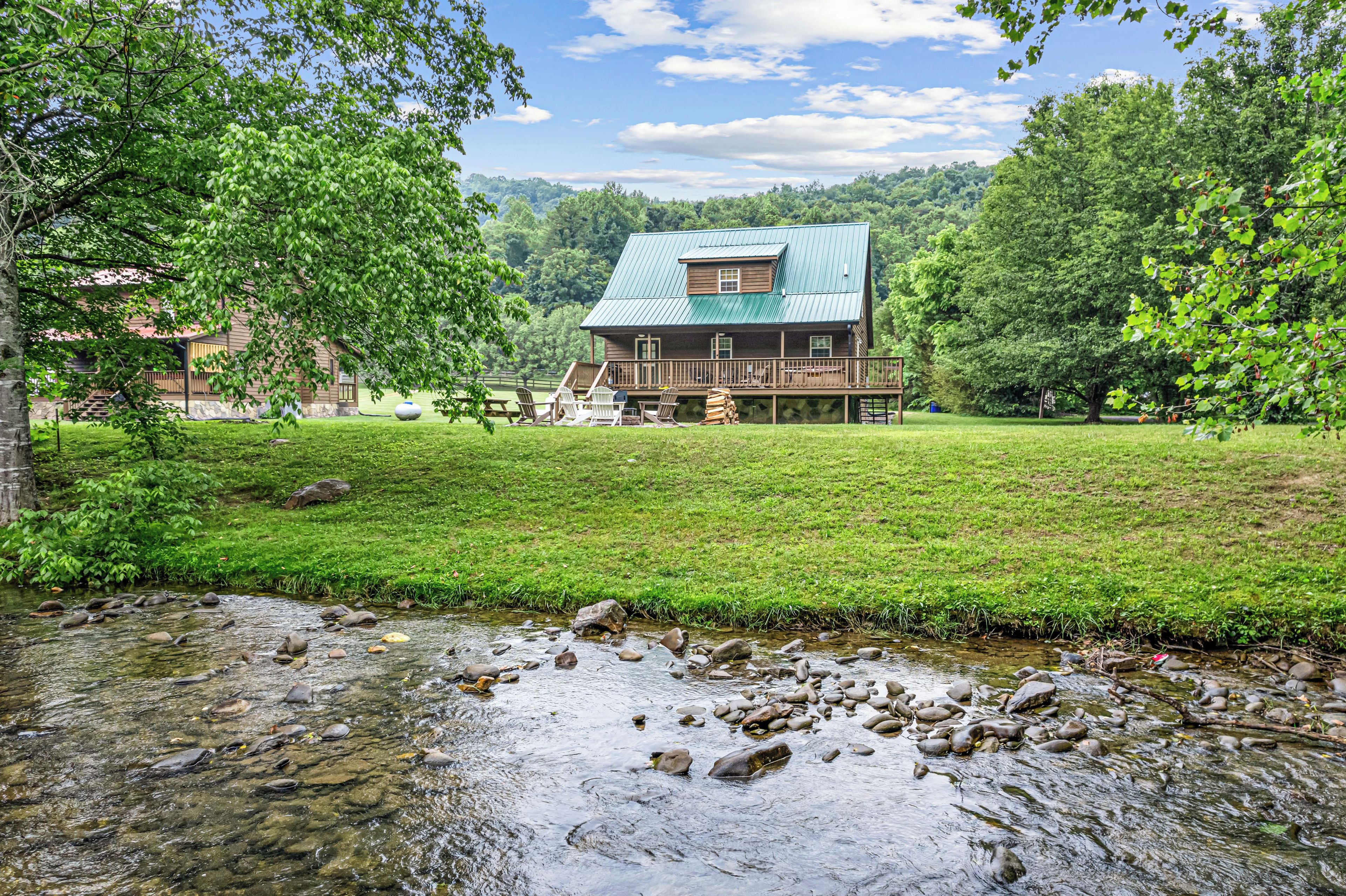 A cabin with a metal roof, surrounded by a grassy area and a stream.