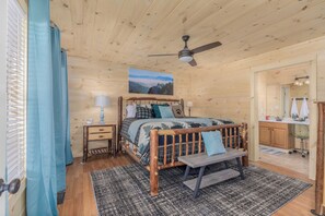 Un dormitorio de cabaña de madera con una cama, mesitas de noche, un ventilador de techo y vistas a las montañas a través de una ventana.