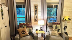 Una habitación con decoración rústica, dos sillones de cuero, una mesita de noche con lámpara, y vistas a un bosque a través de amplios ventanales.