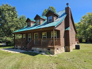 A two-story wooden cabin with a porch, stone foundation, and a green metal roof.