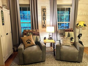 Una habitación con decoración rústica, dos sillones de cuero, una mesita de noche con lámpara, y vistas a un bosque a través de amplios ventanales.